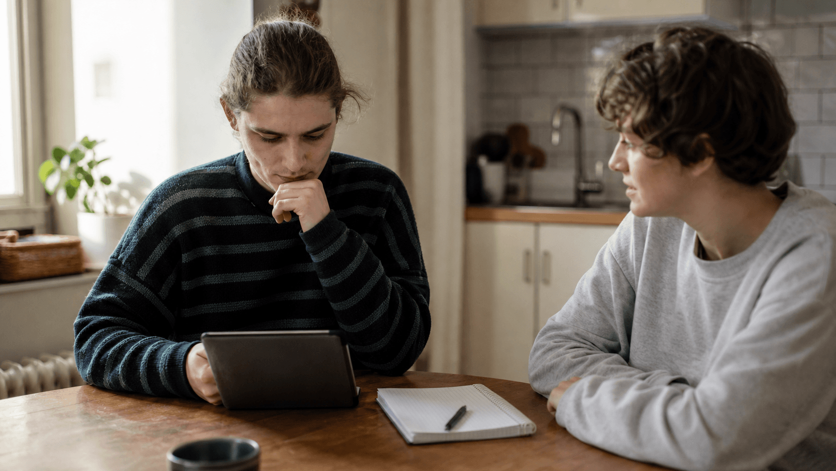 An autistic young adult considers a tablet at home while a friend listens across the table