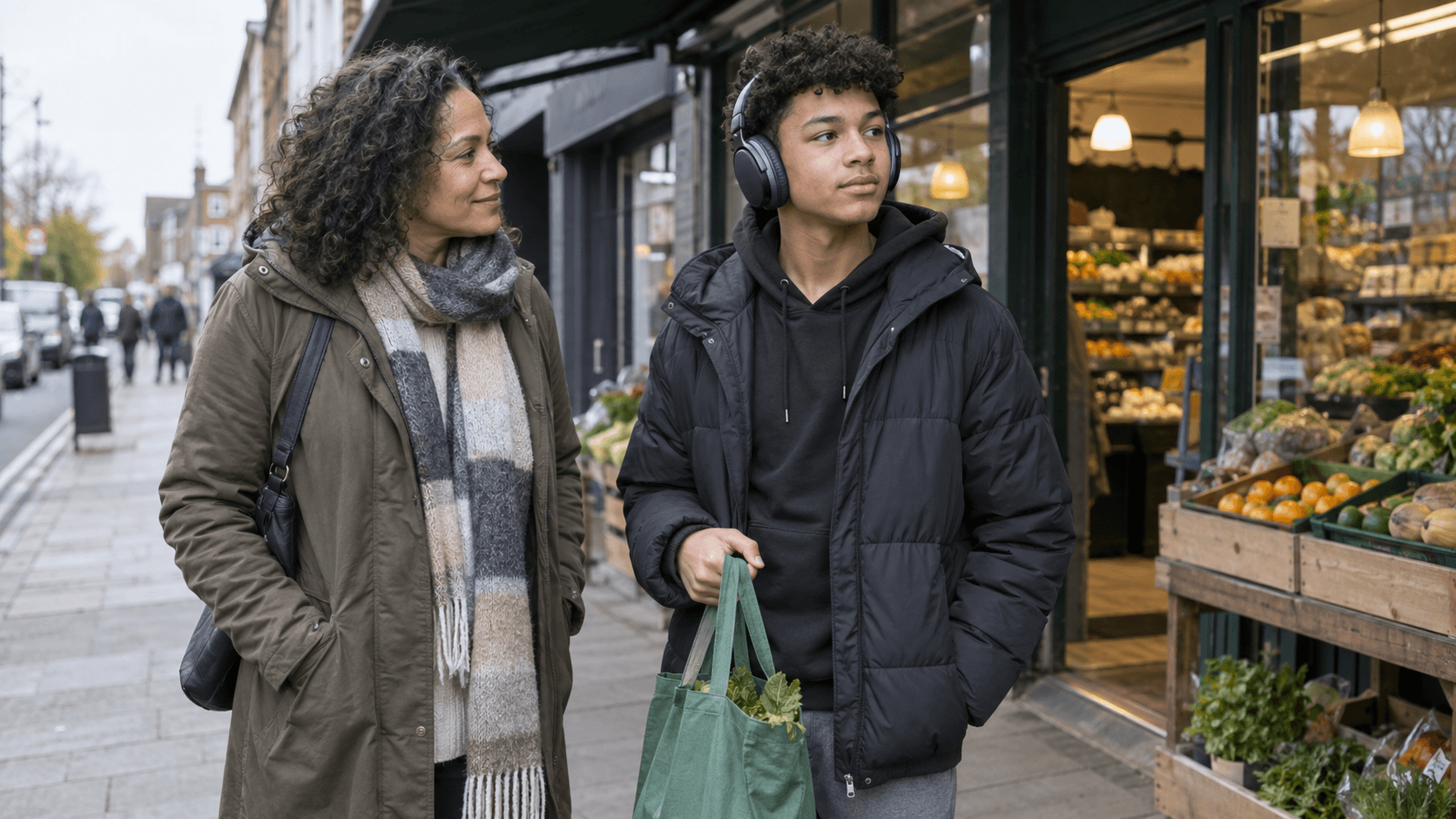 A teenager wearing headphones stands with a parent outside a small grocery shop, holding a reusable shopping bag