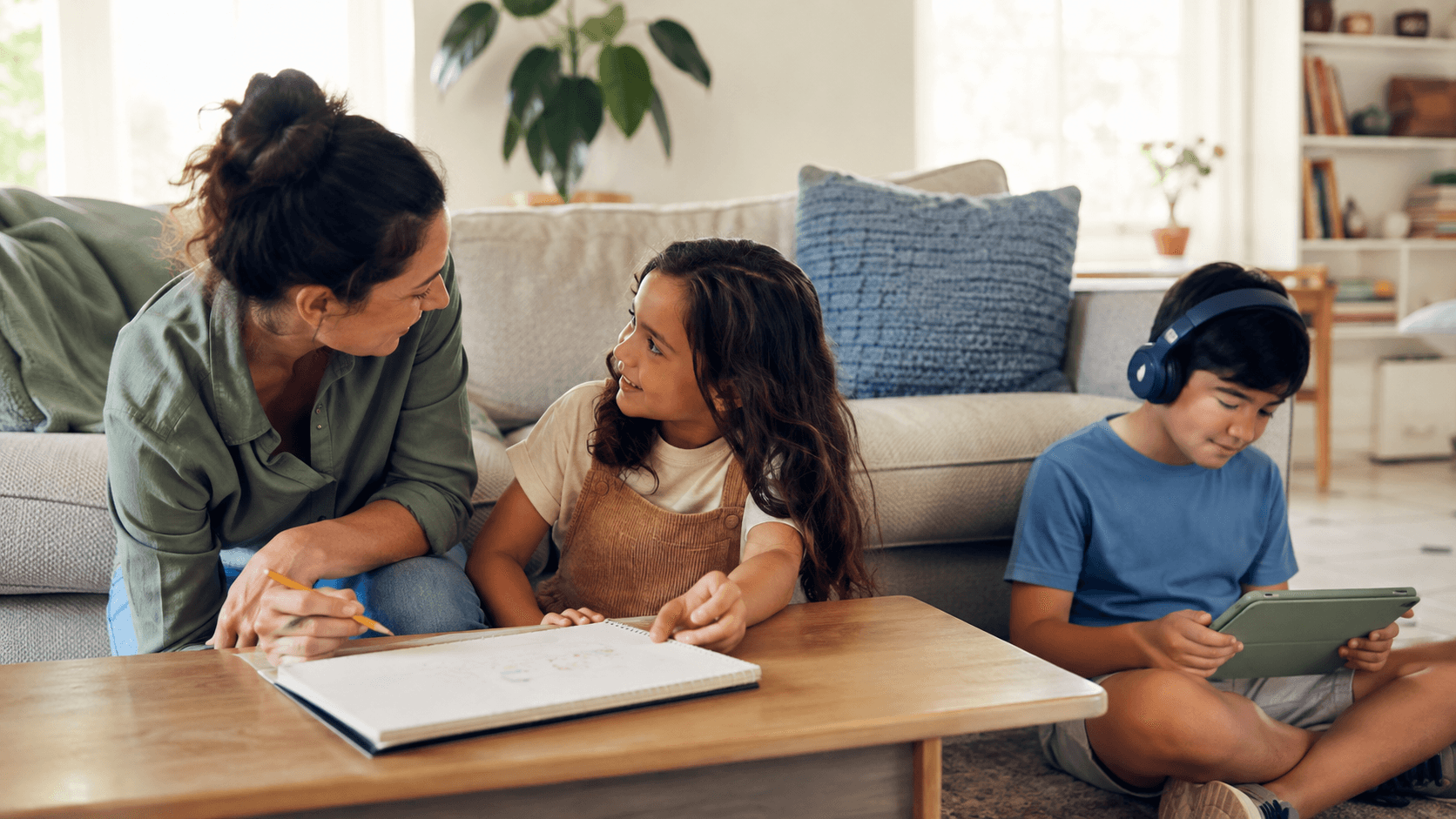 A parent listens to one child over a drawing notebook while their sibling happily uses a tablet nearby in a calm living room