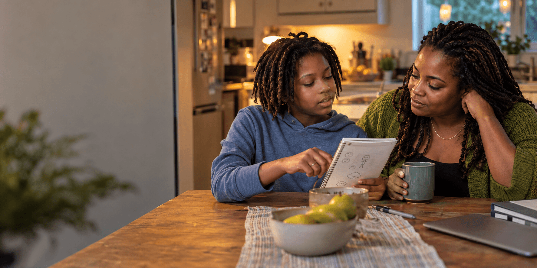 A parent listens while their child points to a notebook at a kitchen table